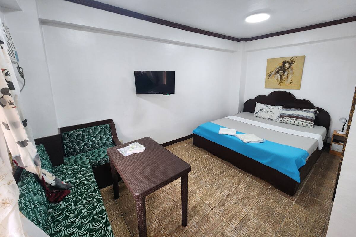 A hotel room with a bed, a small table, a chair, a TV, and a painting on the wall.