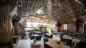 A restaurant with bamboo walls, multiple tables and chairs, a pool table, and a ceiling fan. - MB's Garden Inn (Lapu-Lapu)