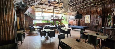 Un restaurante con paredes de bambú, varias mesas y sillas, una mesa de billar y un ventilador de techo.