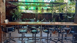 A restaurant with a long wooden bar, black bar stools, and a view of tropical plants through a glass enclosure. - MB's Garden Inn (Lapu-Lapu)