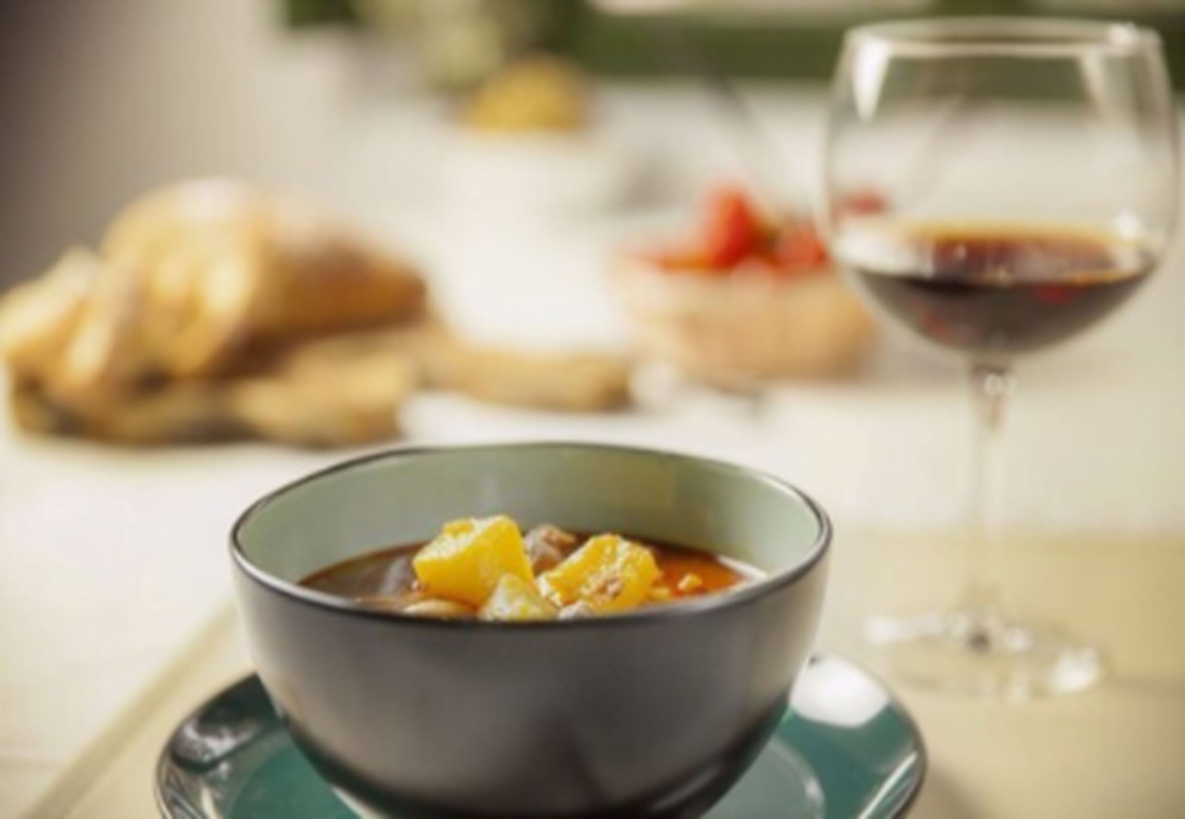 Un tazón de sopa acompañado de una copa de vino tinto y pan al fondo.