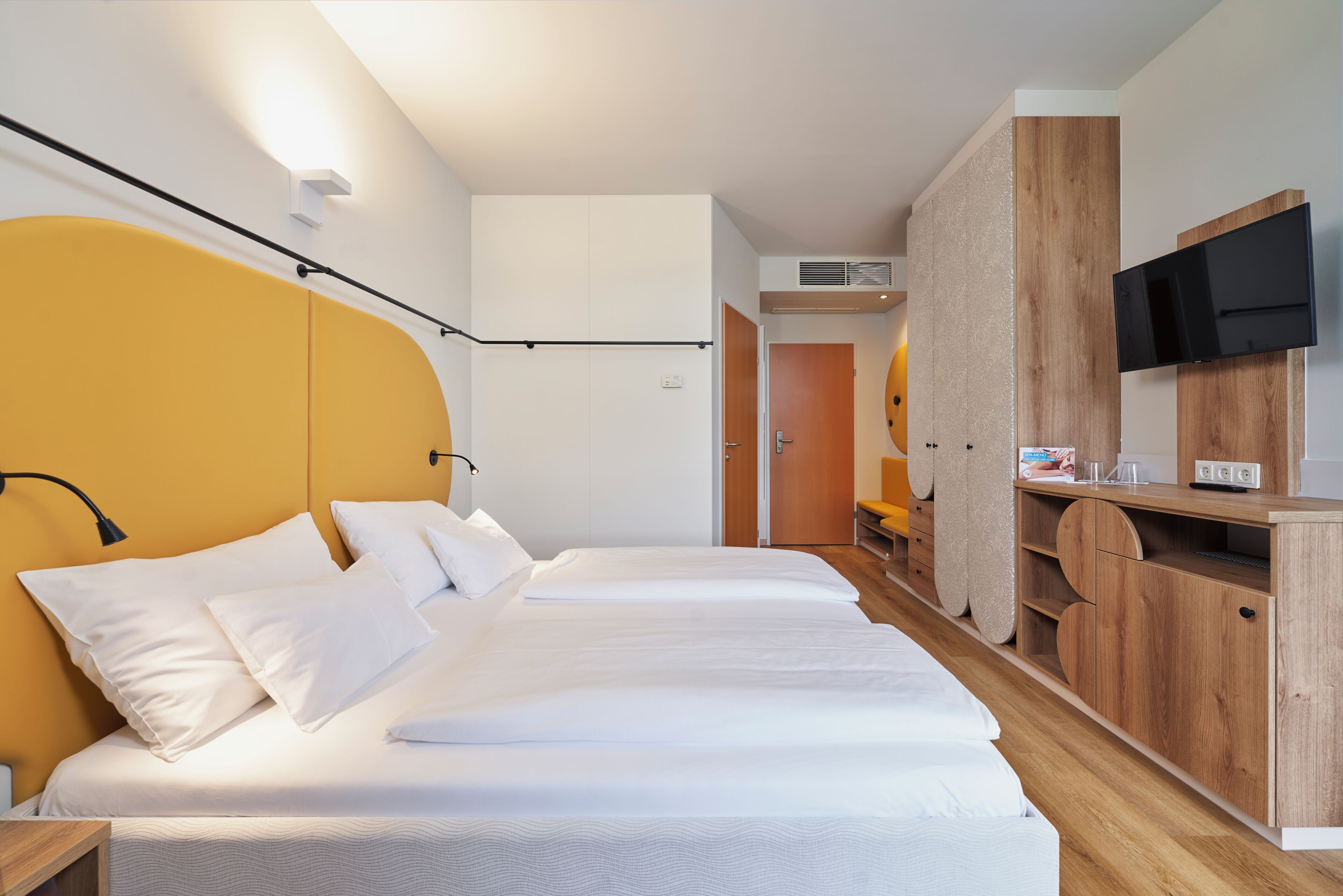 Habitación de hotel con cama, televisión, mueble de madera y puerta que da a otra habitación.