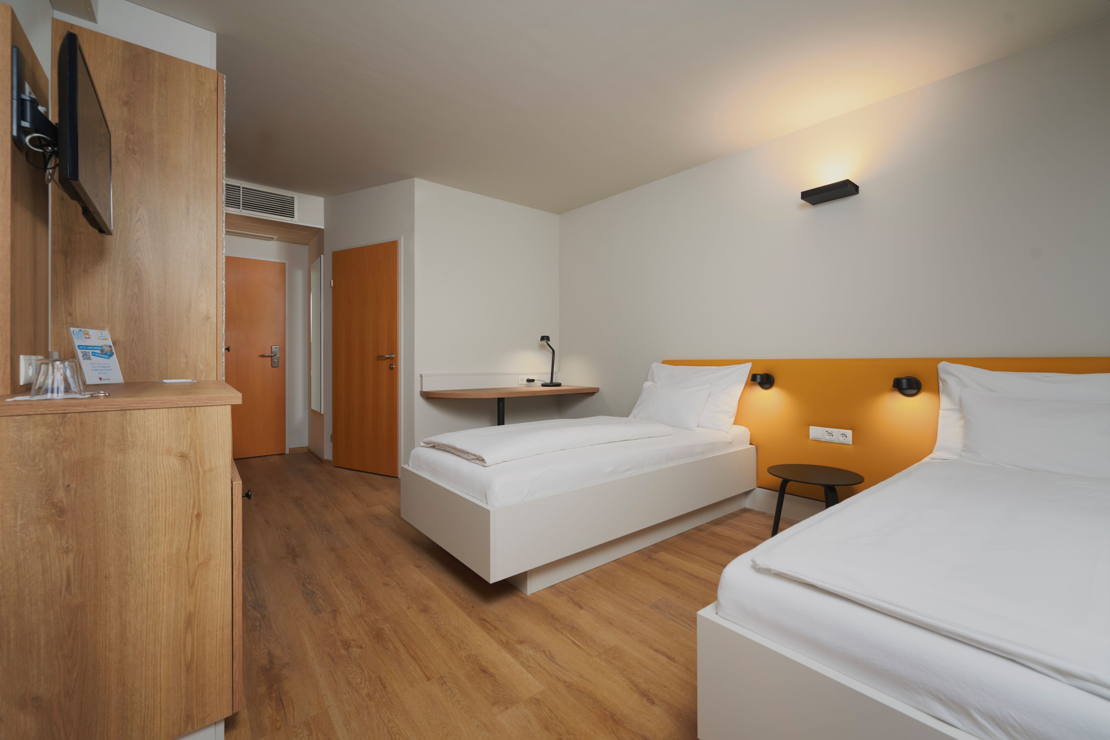 Quarto de hotel com duas camas, uma escrivaninha de madeira, uma mesa pequena e televisão na parede.