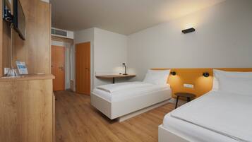 Quarto de hotel com duas camas, uma escrivaninha de madeira, uma mesa pequena e televisão na parede.