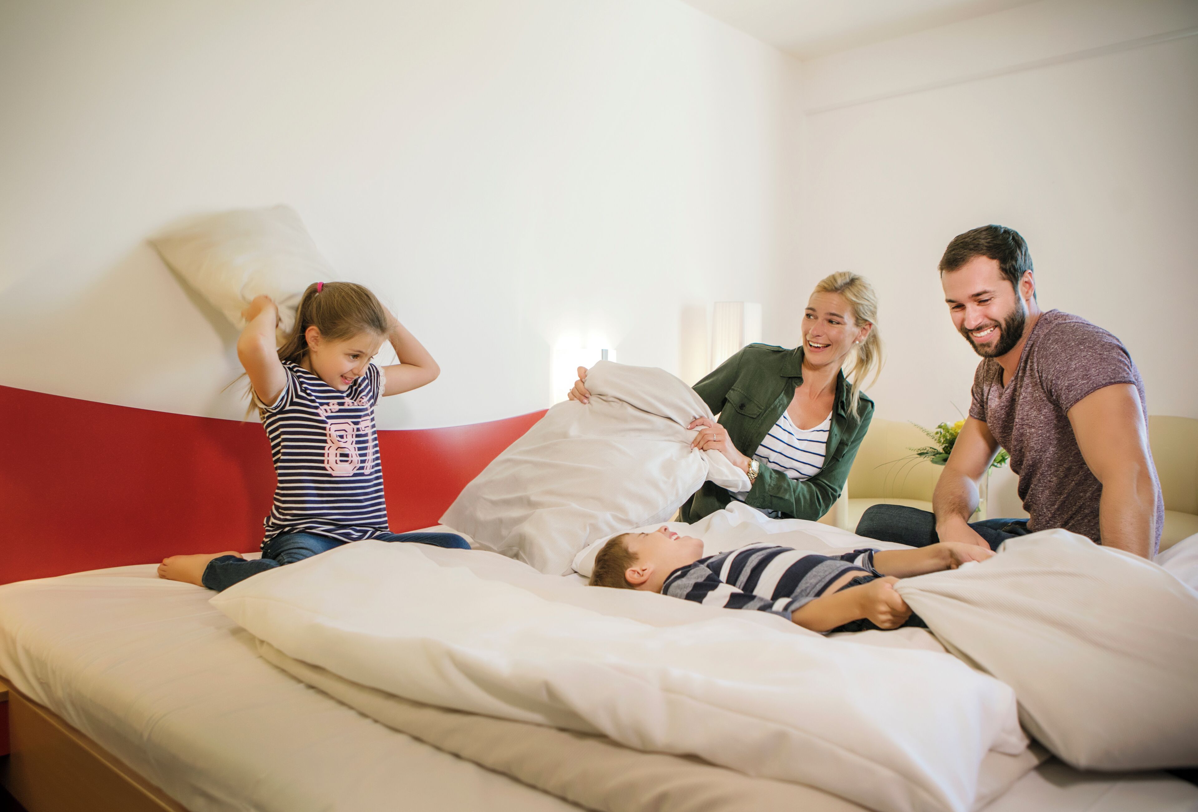 A family in a bedroom with a bed, pillows, and a red headboard.