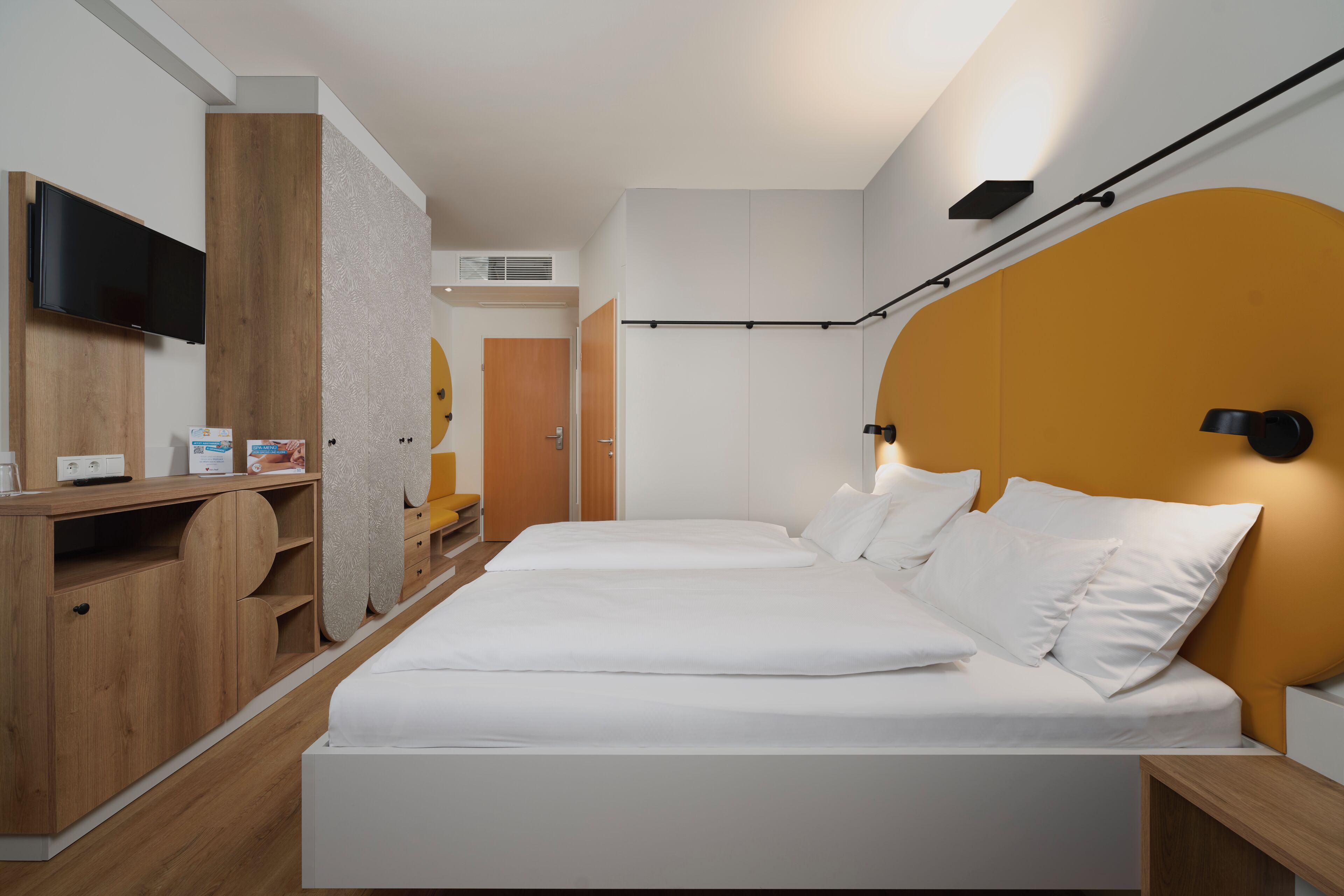 A modern hotel room with a bed, wooden furniture, a flat-screen TV, and a wall-mounted light fixture.