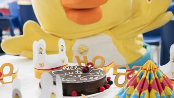 A large yellow duck mascot with a red cap, surrounded by party decorations including a chocolate cake, party hats, and a colorful paper fan.