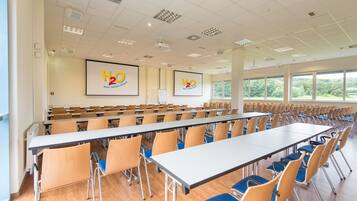 A large conference room with rows of tables and chairs, large windows, and two screens displaying 'H2O Hotel-Themed Resort'.