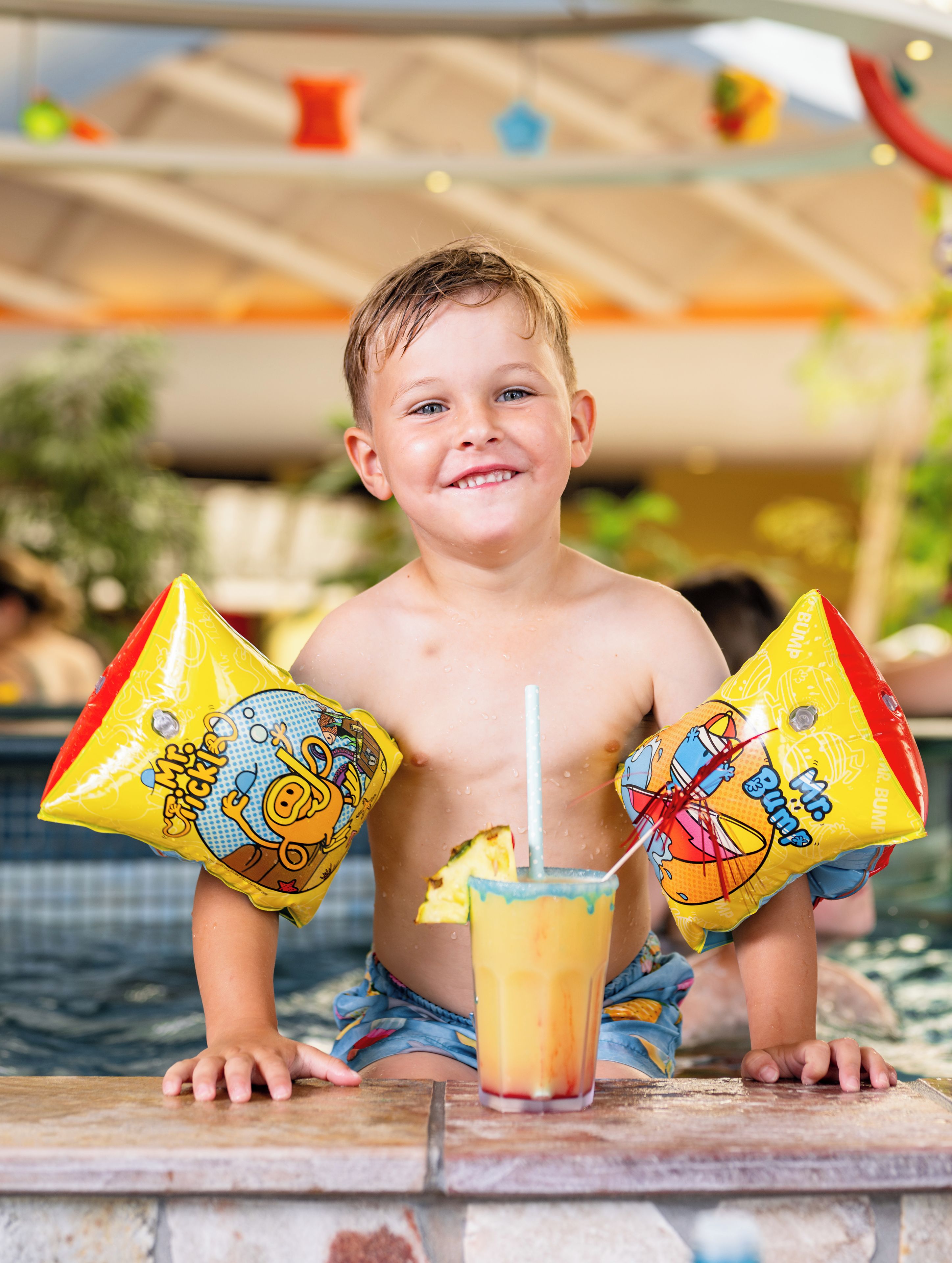 Niño sentado junto a una piscina con dos pelotas de playa Mr. Bubbles y una bebida.
