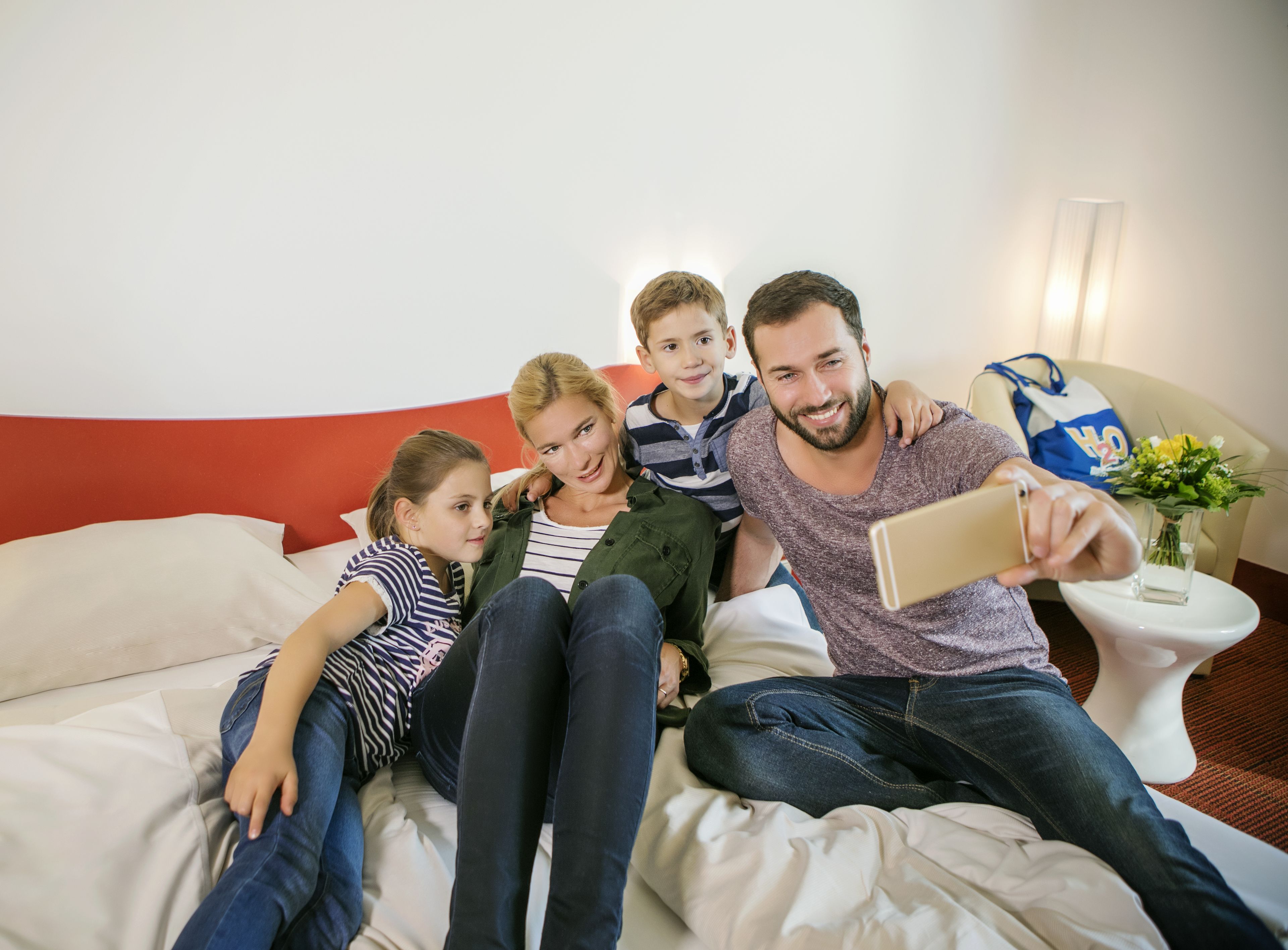 Una familia de cuatro personas en una cama con cabecera roja, una mesita de noche blanca con un florero y un bolso azul sobre la mesita.