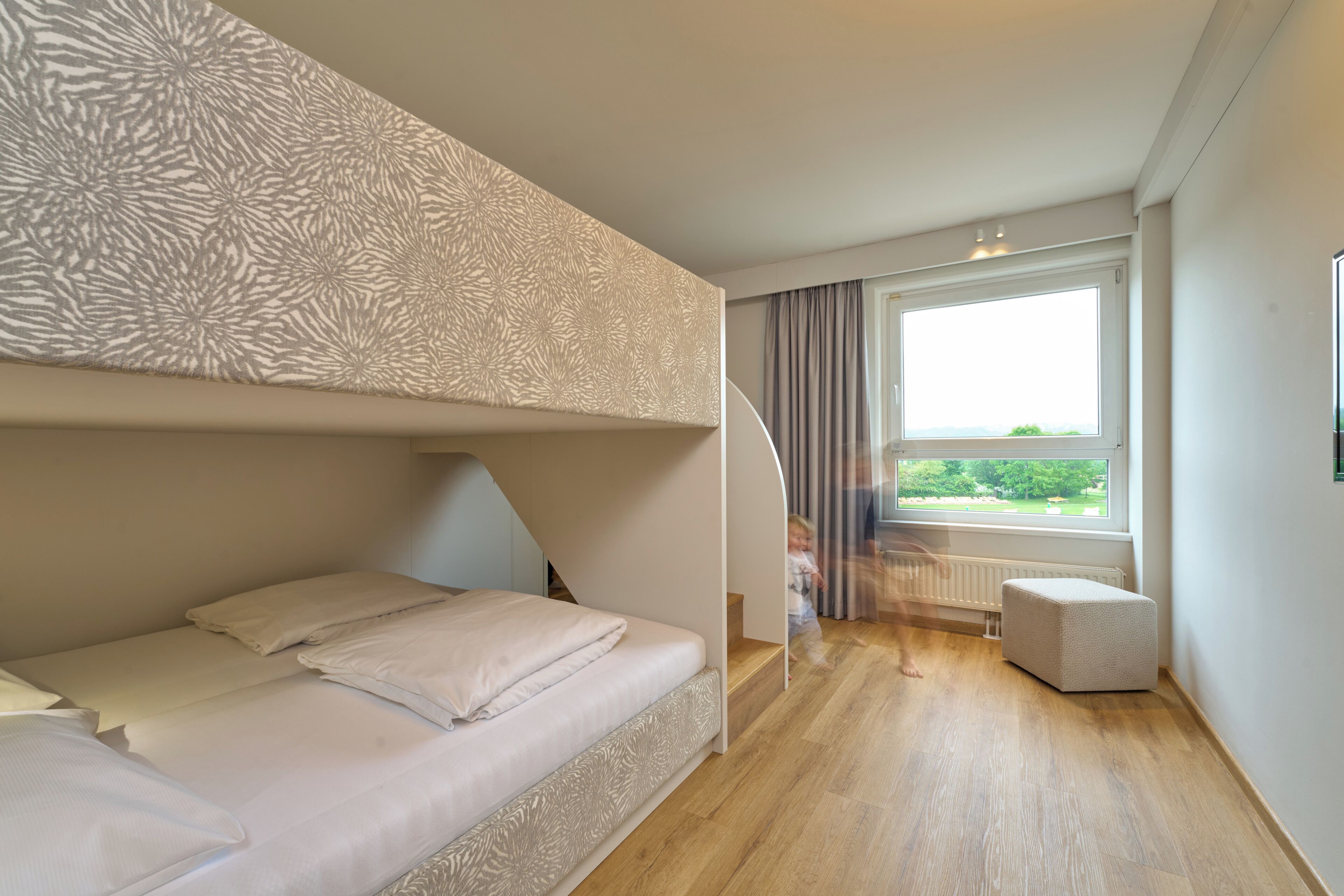 A bedroom with a bunk bed, a window with a view, a small ottoman, and a radiator.
