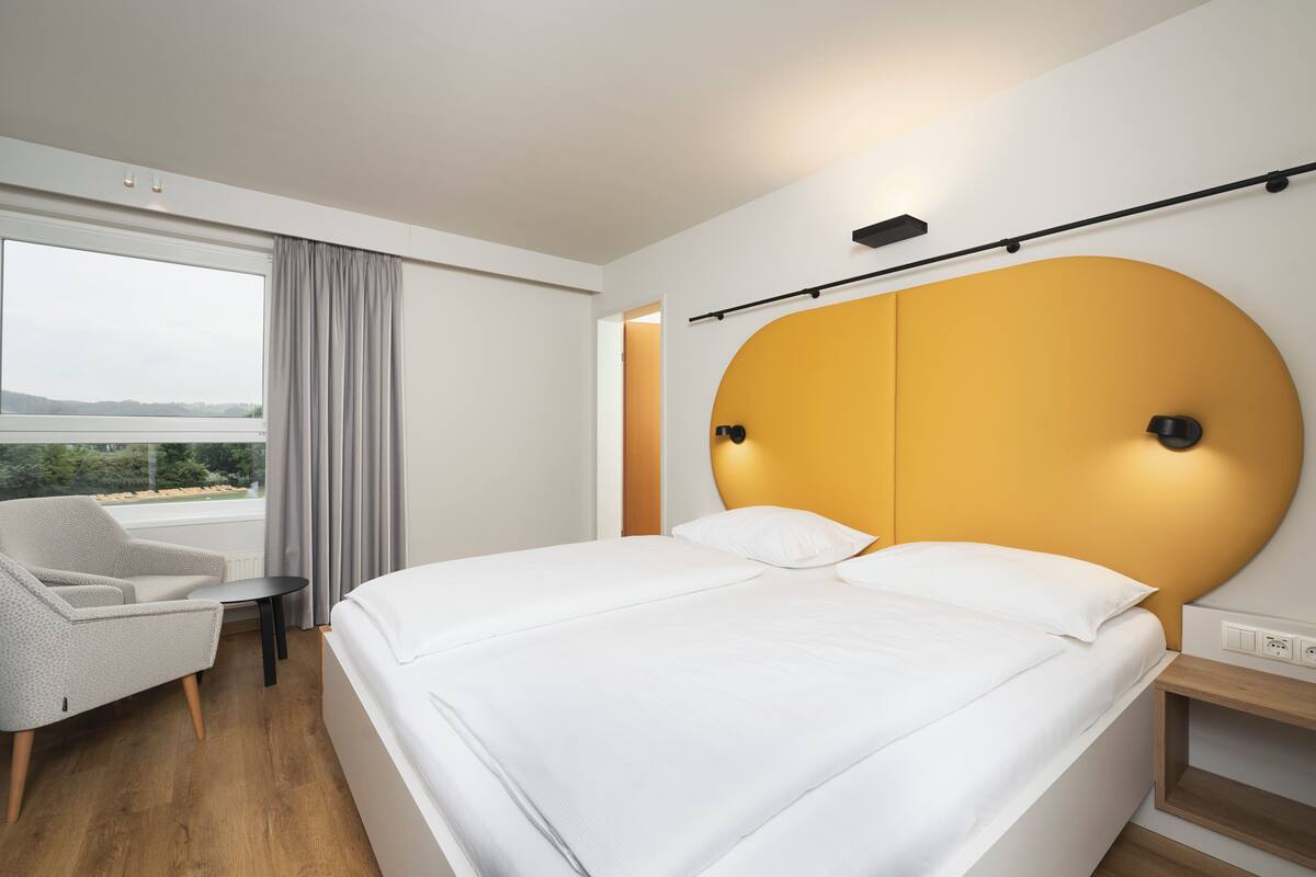 Een moderne hotelkamer met een groot bed, een zithoek met een stoel, een klein tafeltje en uitzicht op het groen.