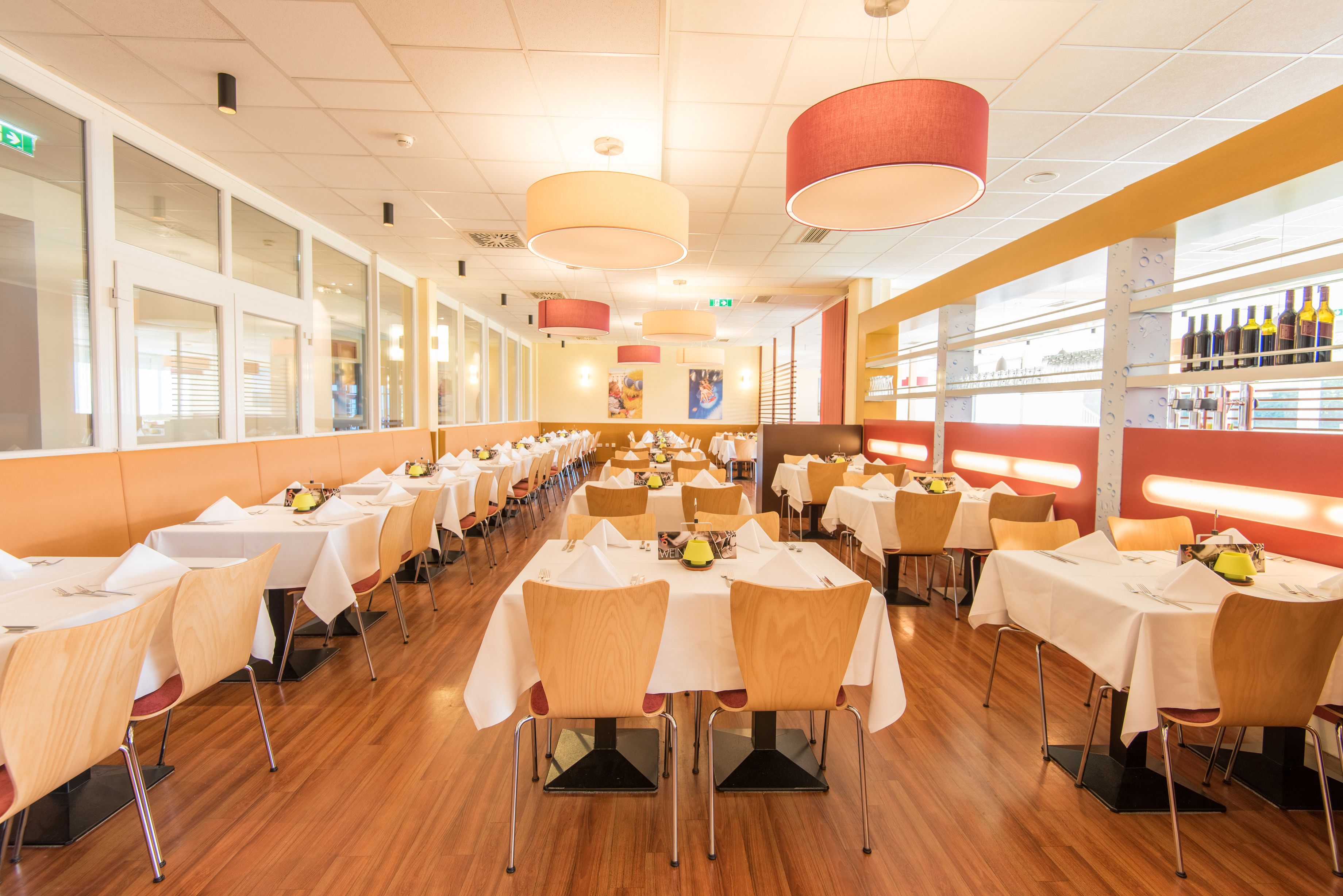 A spacious restaurant with multiple tables and chairs, a long counter with a wine display, and large windows along one side.