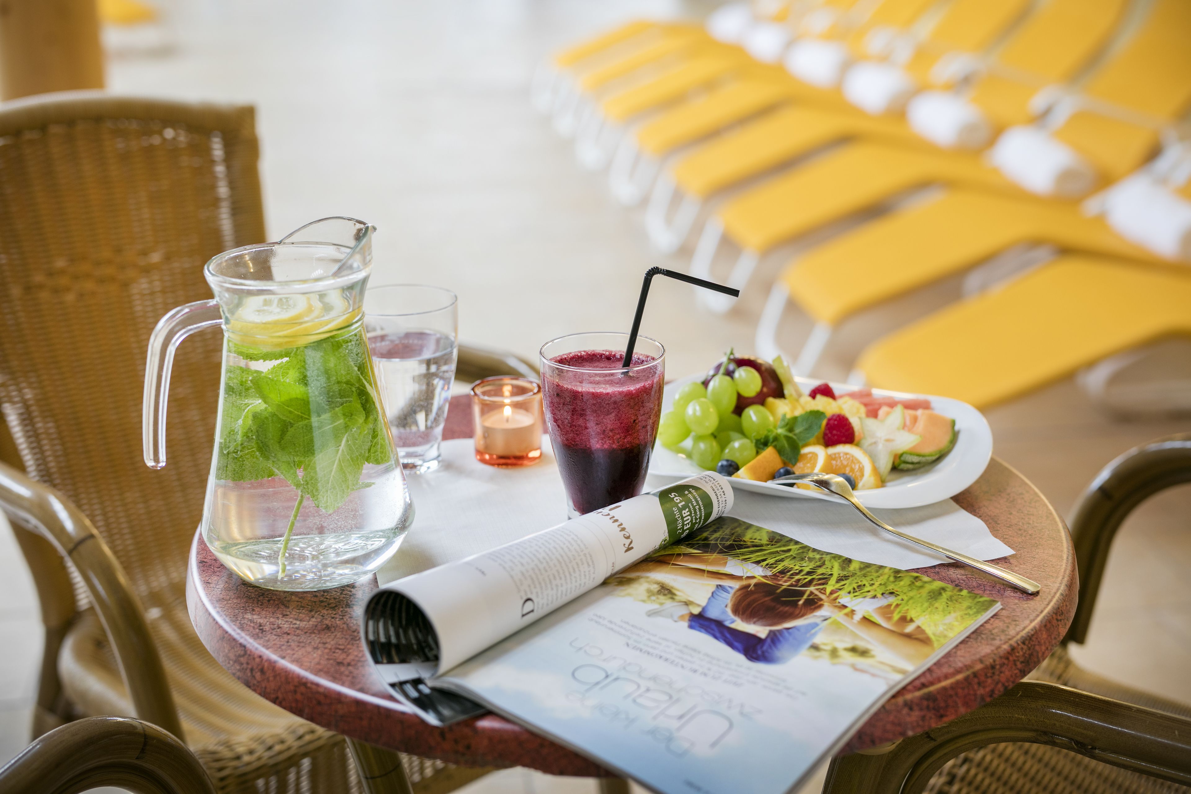 A table with a pitcher of water, a glass of juice, a plate of fruit, a candle, and a magazine.