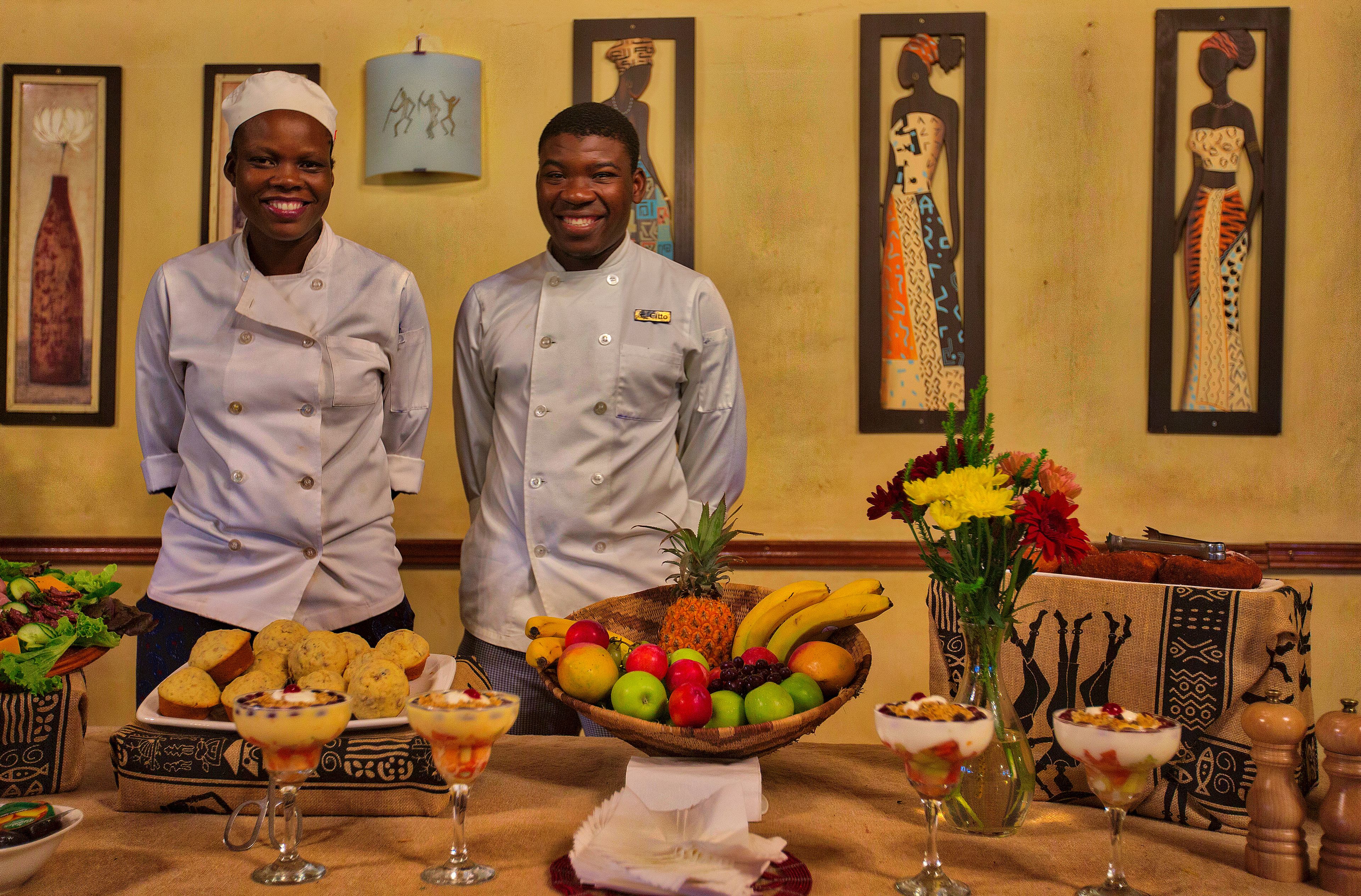 Two chefs standing behind a table with a fruit bowl, desserts, and a vase of flowers.