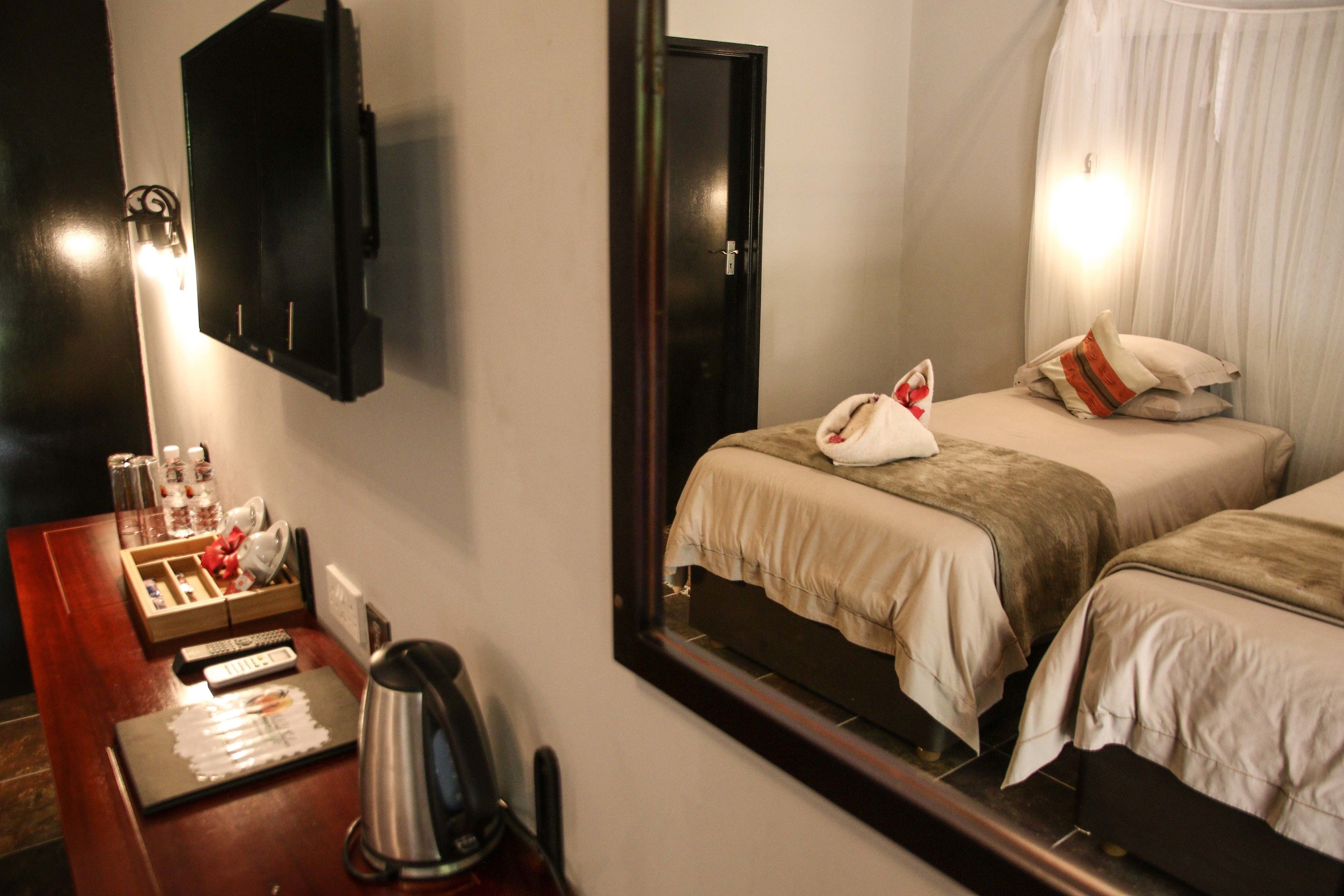 Quarto de hotel com duas camas, uma mesa com chaleira, minibar e um espelho refletindo o aposento.