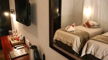 Quarto de hotel com duas camas, uma mesa com chaleira, minibar e um espelho refletindo o aposento.
