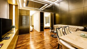 Moderni hotellihuone, jossa on puulattia, taulutelevisio, ruokapöytä tuoleineen ja kattokruunu.