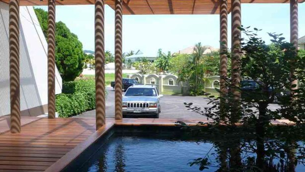 Un espacio exterior techado con piscina, pilares de madera y vistas a un estacionamiento y vegetación.