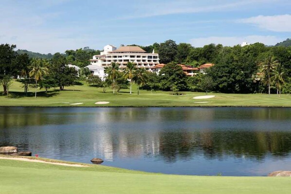 A golf course with a large building in the background, surrounded by greenery and a body of water. - The Green Golf Residence Condominium (Kathu)