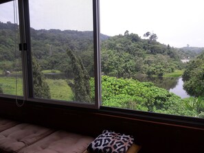 A room with a large window overlooking a lush landscape with a river and dense greenery. - The Green Golf Residence Condominium (Kathu)