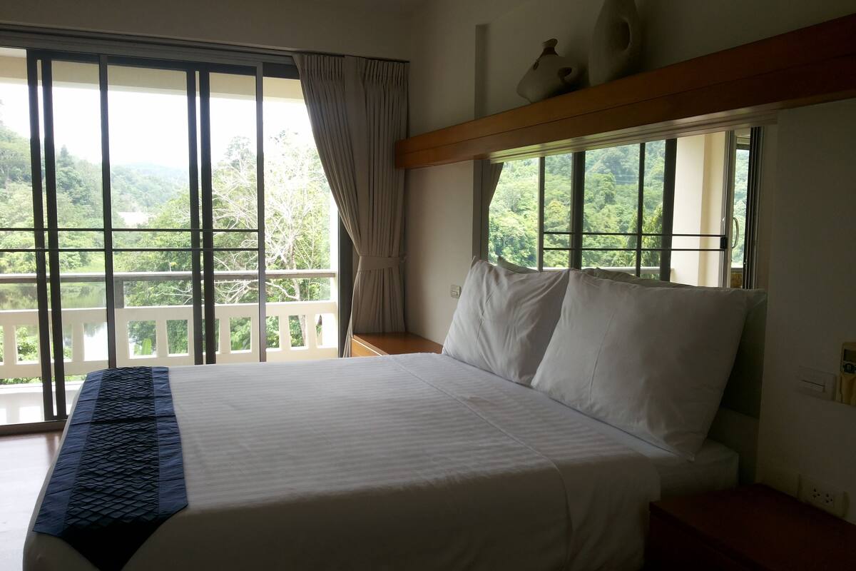 A hotel room with a large bed, a view of a lake and forest through the window, and a wooden nightstand.