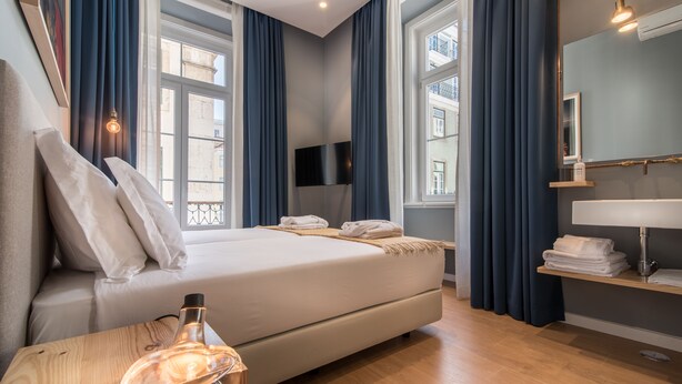 Una habitación de hotel moderna con una cama grande, una mesita de noche con lámpara, un espejo y una ventana con cortinas.
