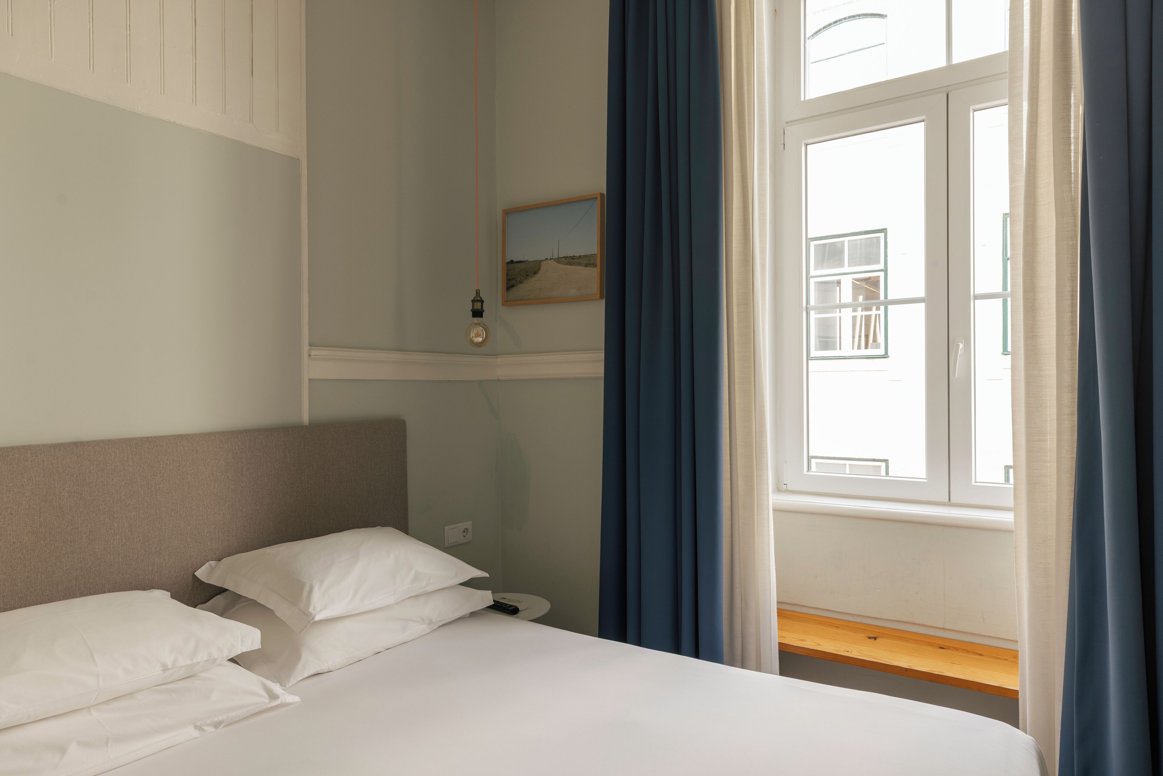 A hotel room with a bed, a bedside table, a nightstand, a window with curtains, and a wall-mounted picture.