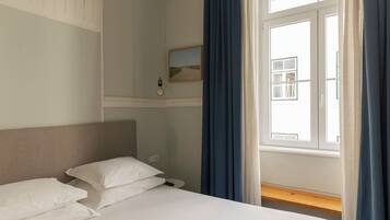 A hotel room with a bed, a bedside table, a nightstand, a window with curtains, and a wall-mounted picture.