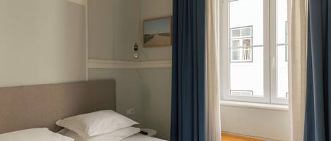 A hotel room with a bed, a bedside table, a nightstand, a window with curtains, and a wall-mounted picture.