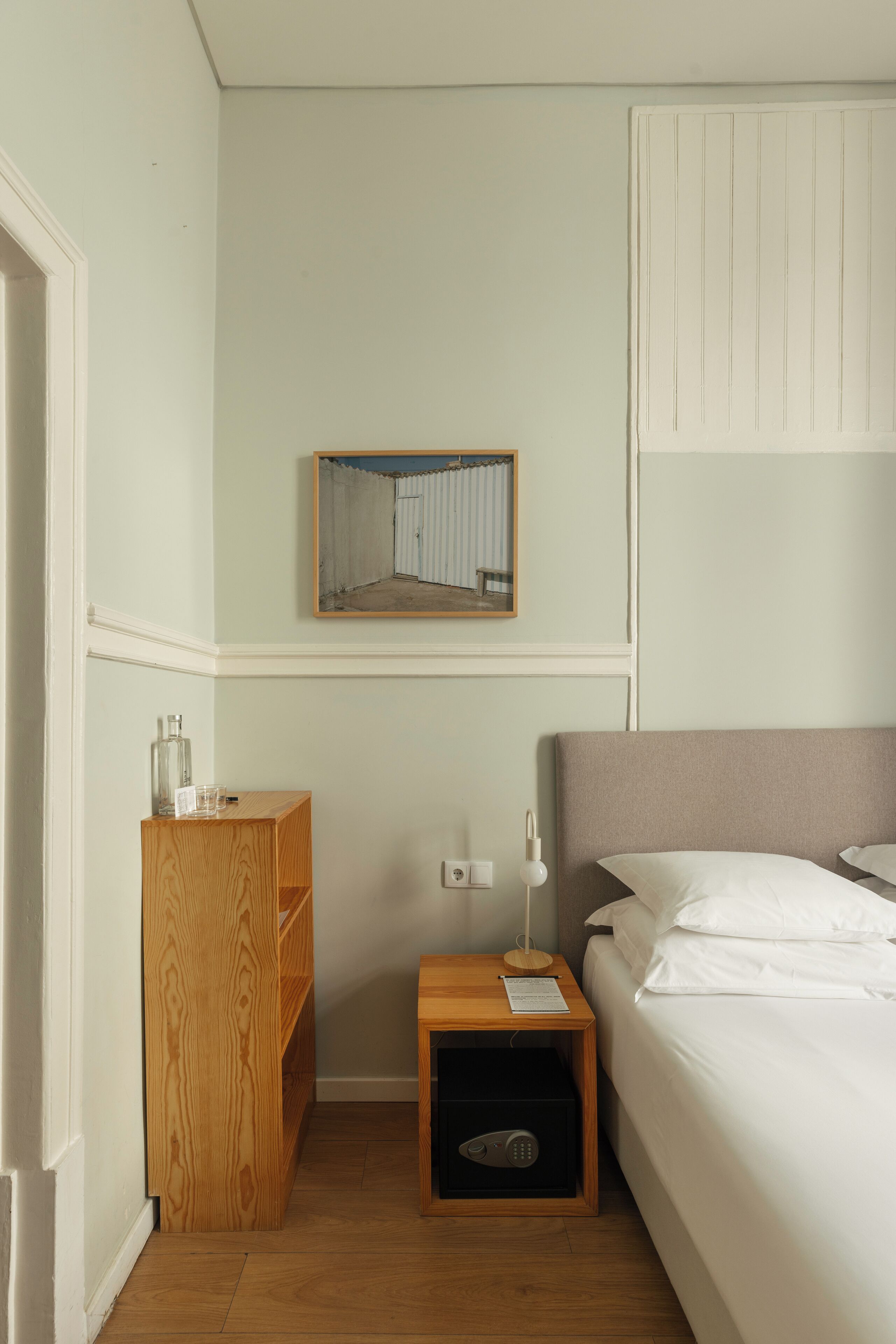 A small hotel room with a bed, a nightstand, a wooden cabinet, a bedside table, a mirror, and a wall-mounted light.