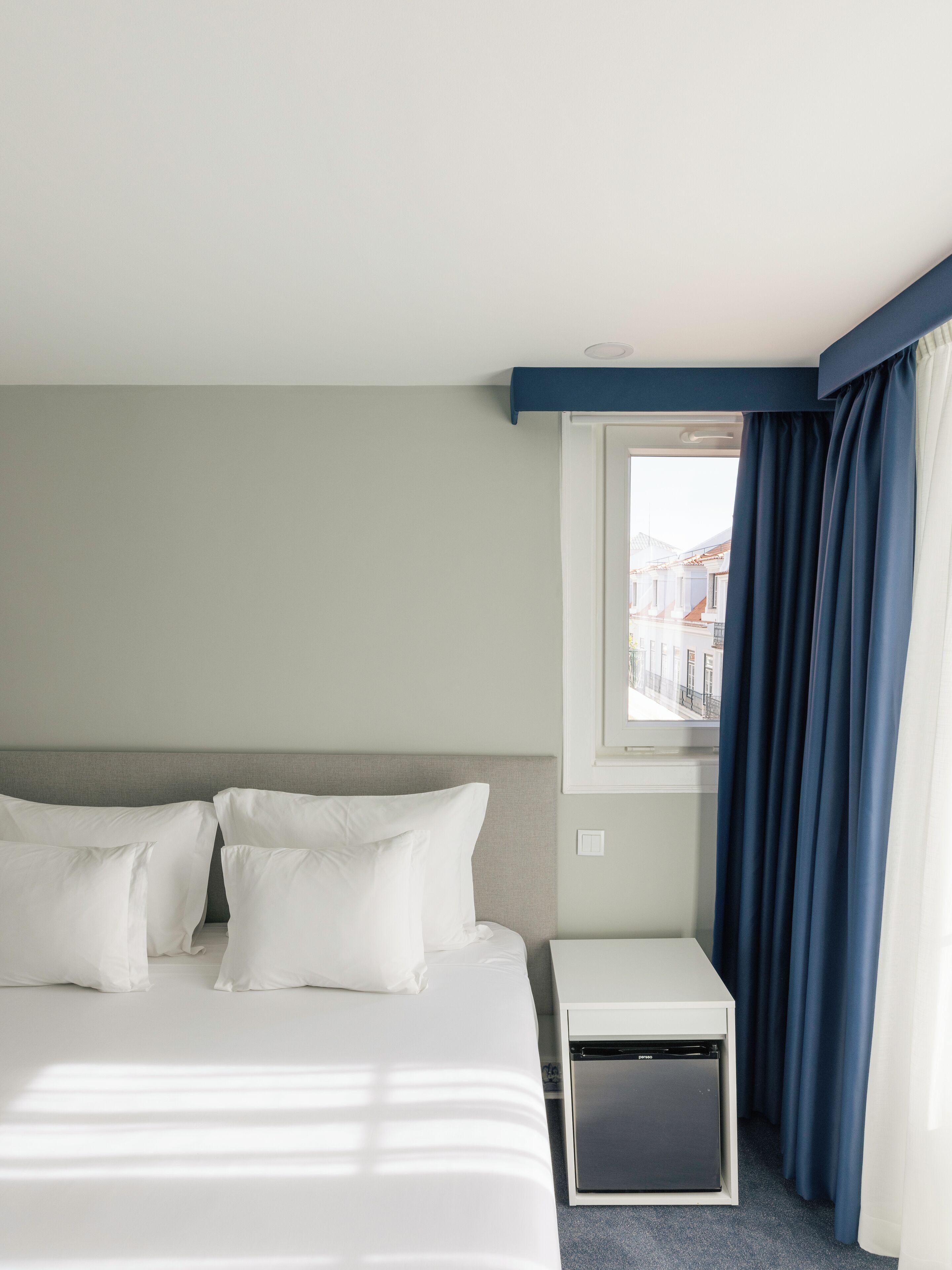 A hotel room with a bed, a small bedside table, a window with curtains, and a view of buildings outside.