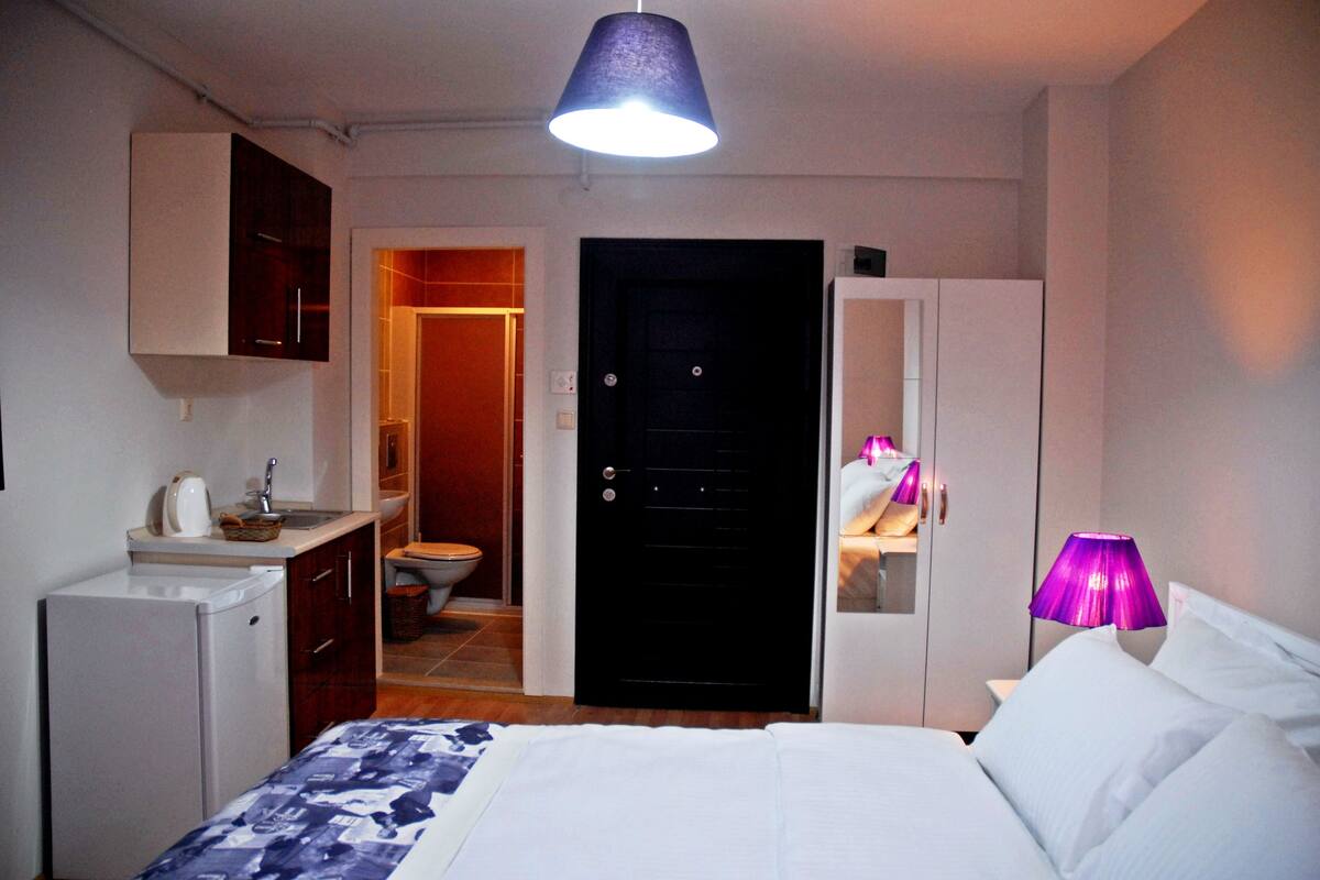 Ein kompaktes Hotelzimmer mit Bett, einer kleinen Küchenzeile, einem Badezimmer und einer Tür zu einem weiteren Raum.