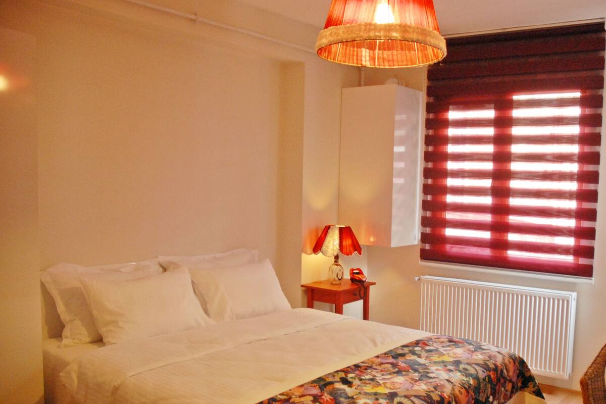 Ein Schlafzimmer mit Bett, Nachttisch mit Lampe, Heizkörper und Fenster mit Jalousien.