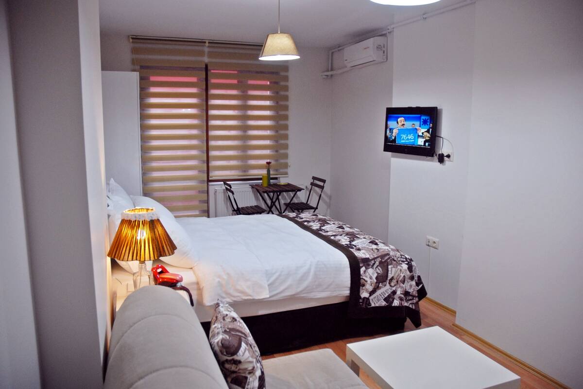 Ein kleines, gut beleuchtetes Schlafzimmer mit Bett, Nachttisch, Lampe und einem an der Wand montierten Fernseher.