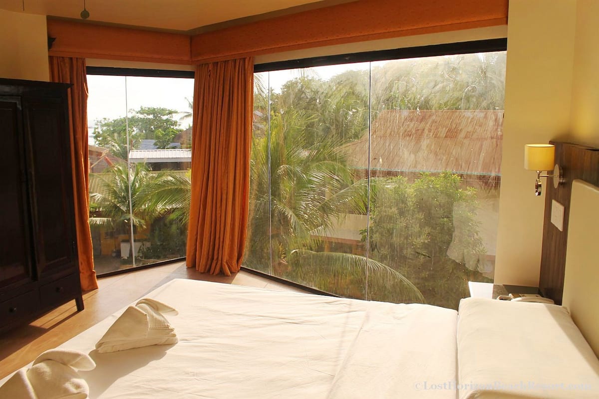 A hotel room with a large sliding glass door leading to a balcony with a view of palm trees and a house. There is a bed with white linens and a bedside table with a lamp.
