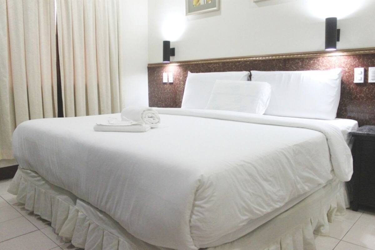 A neatly made bed with white linens, a bedside table with a lamp, and a wall-mounted light fixture.