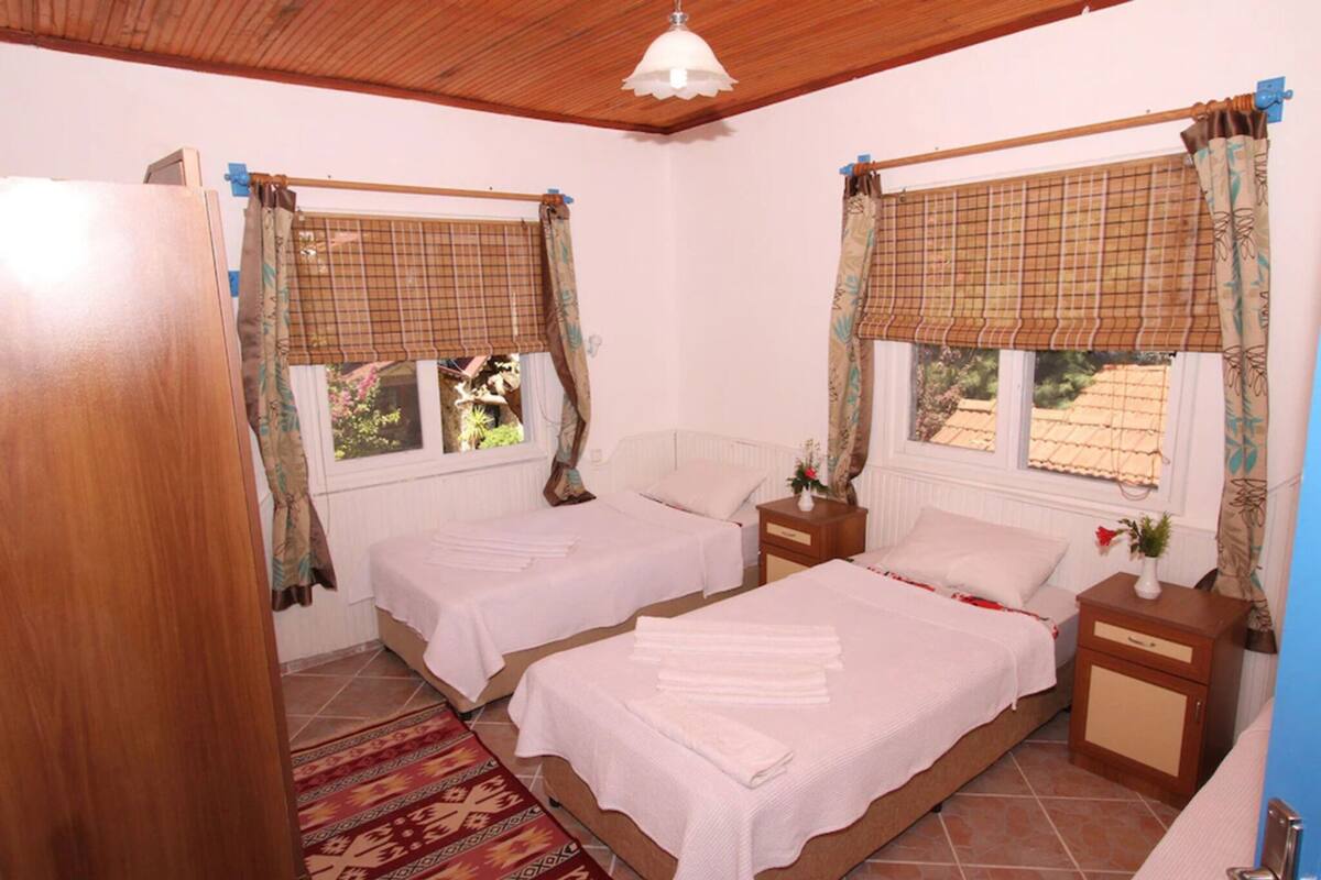 Habitación con dos camas, techo de madera, ventana con cortina, mesita de noche y alfombra estampada.