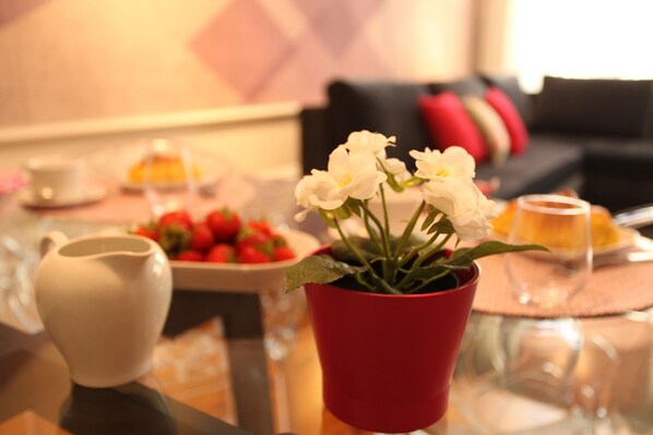 Une table à manger avec un pichet blanc, un pot rouge contenant des fleurs blanches, un saladier de fraises et un verre d’eau.