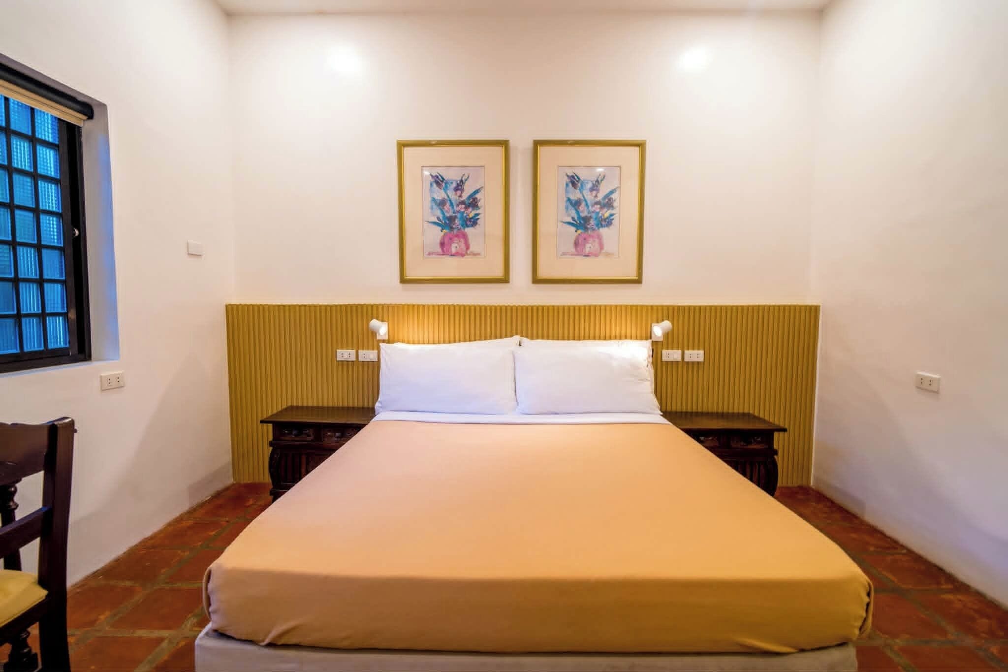 A hotel room with a bed, two bedside tables, a chair, a window, and two framed artworks on the wall.