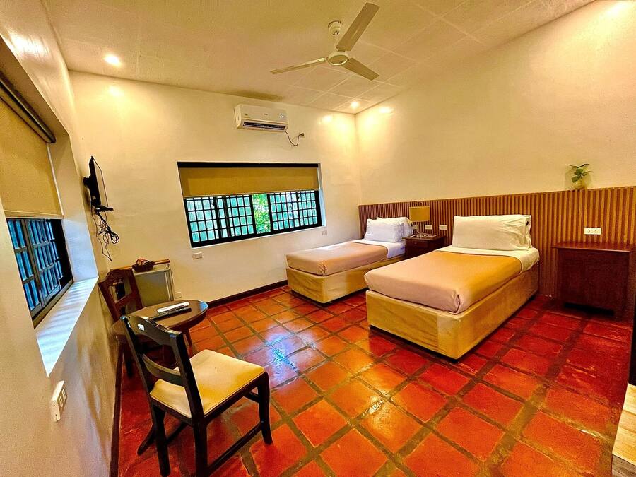 Une chambre d’hôtel avec deux lits, un bureau, une chaise, une fenêtre donnant sur un paysage agréable, un ventilateur de plafond et un appareil de climatisation.