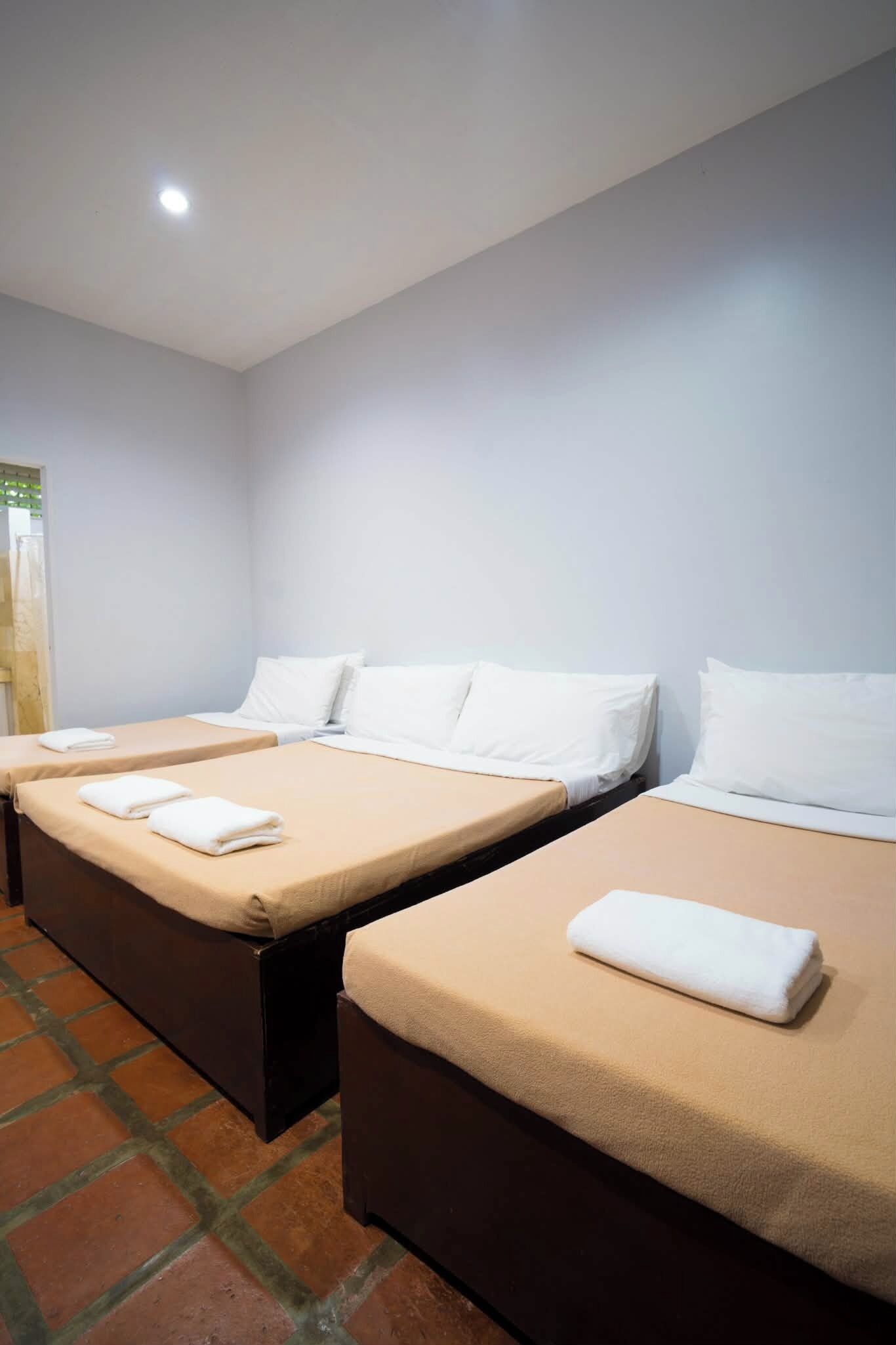 Ein Hotelzimmer mit drei nebeneinander liegenden Betten, jedes mit einem weißen Kopfkissen und einem gefalteten weißen Handtuch.