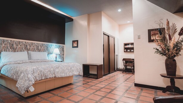 Een hotelkamer met een bed, een nachtkastje, een lamp, een kleine tafel en een vaas met gedroogde planten.