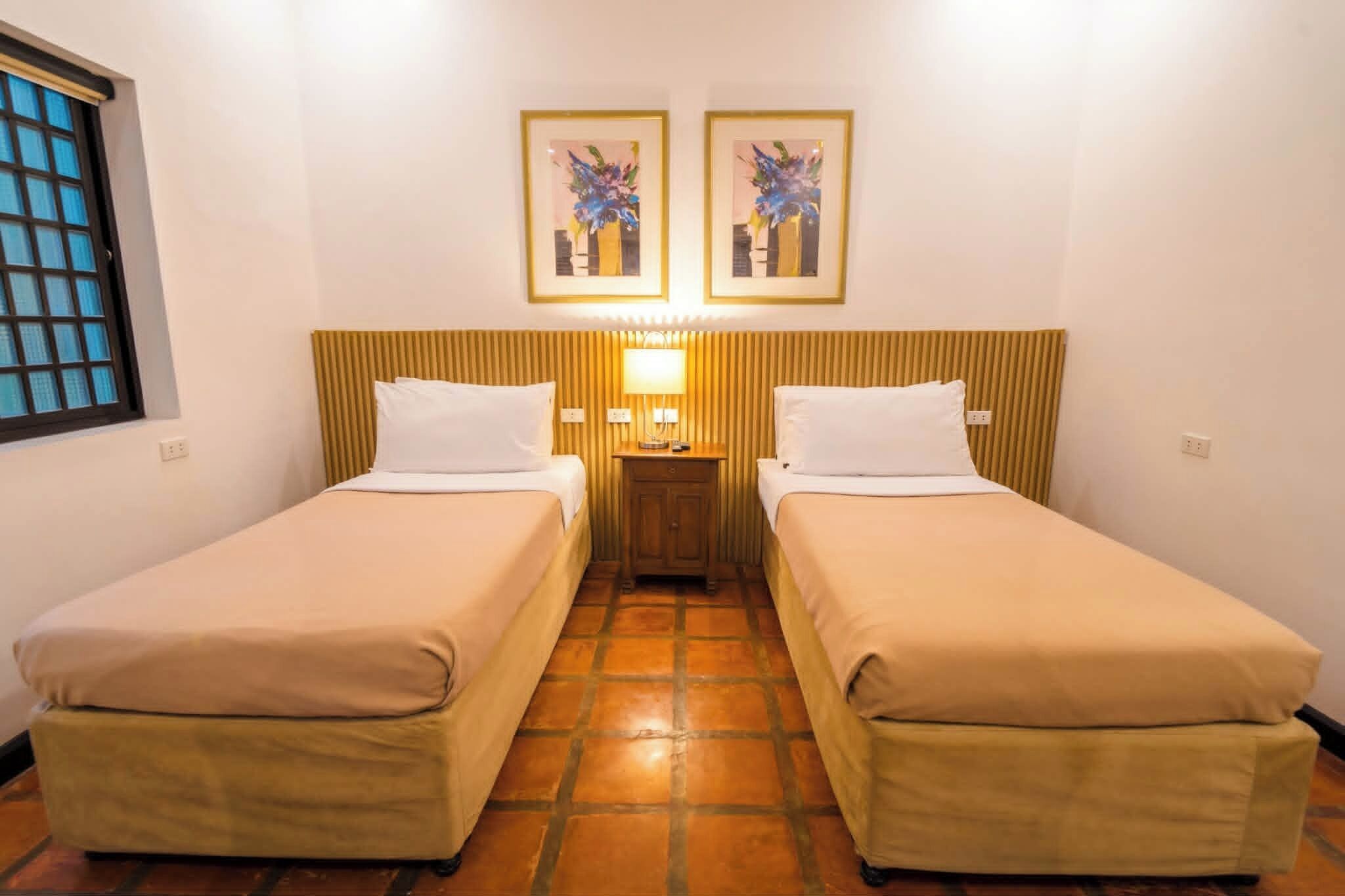 Habitación de hotel con dos camas individuales, una mesita de noche con lámpara y dos cuadros enmarcados con motivos florales en la pared.