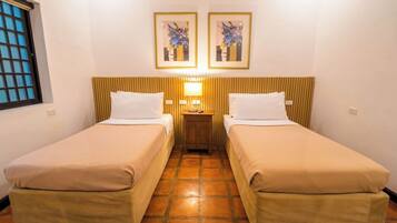 Habitación de hotel con dos camas individuales, una mesita de noche con lámpara y dos cuadros enmarcados con motivos florales en la pared.