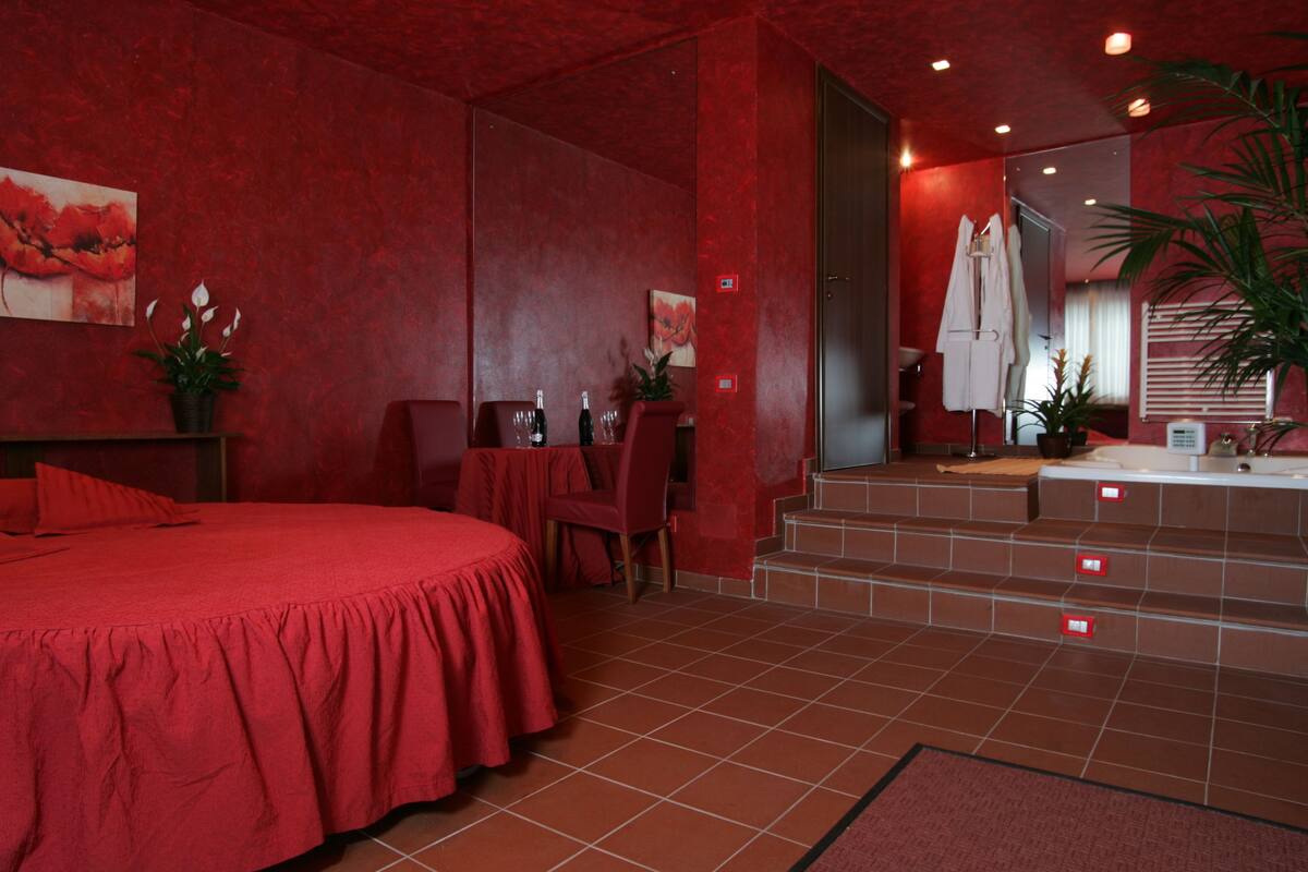 A red-themed hotel room with a large bed, a small table with chairs, a bathroom with a mirror, and a staircase leading to an upper level.