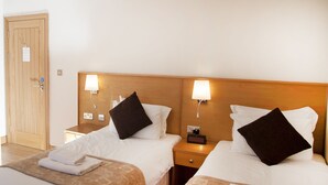 A hotel room with two beds, each with a wooden headboard, white bedding, and a black pillow. There are bedside tables with lamps and a small clock. A wooden door is visible in the background. - Somerfield Lodge (Swansea)