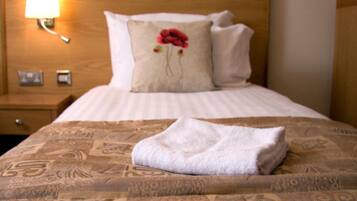 A neatly made bed with a patterned bedspread, a pillow with a red flower, and a folded towel on the bed.