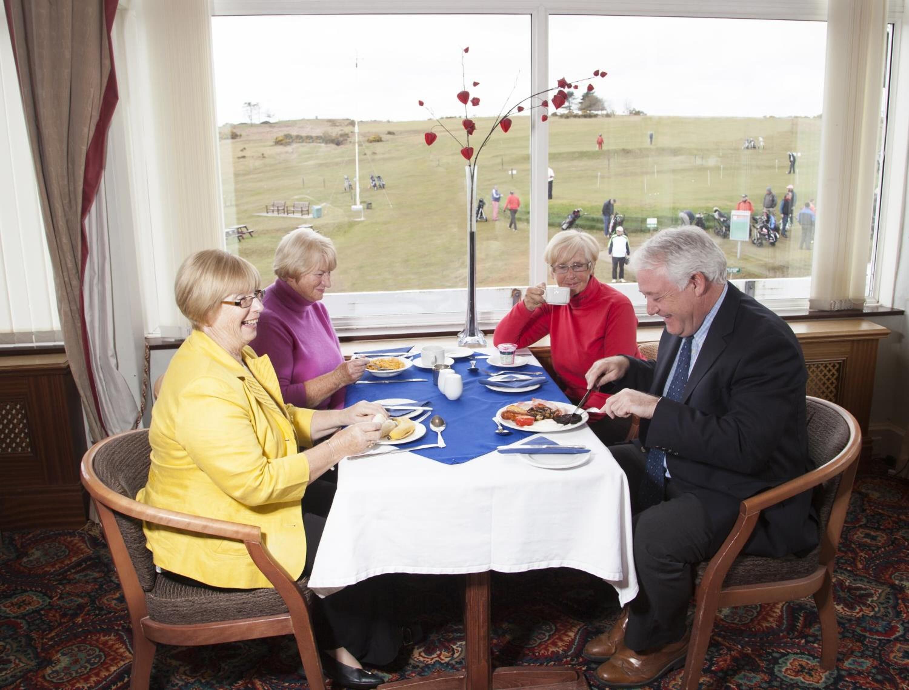 Un grupo de personas cenando en una mesa con vista a un campo verde y gente caminando a lo lejos.