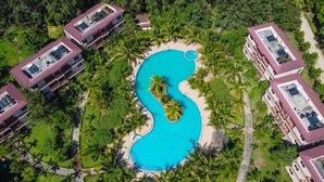 An aerial view of a resort with a central swimming pool, surrounded by lush greenery and palm trees.